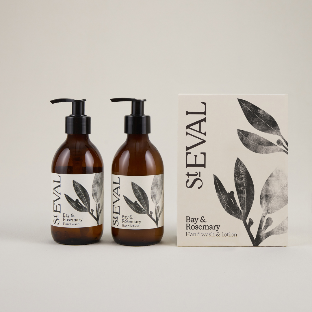 Two brown glass jars with pumps sitting next to a beige box with a foliage pattern on the right hand side on a plain grey background, featuring the branding 'St Eval' in black text.