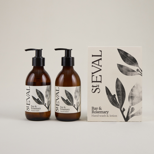 Two brown glass jars with pumps sitting next to a beige box with a foliage pattern on the right hand side on a plain grey background, featuring the branding 'St Eval' in black text.