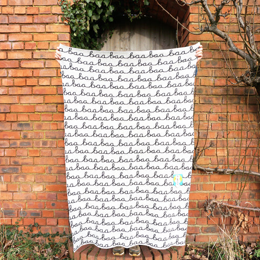 A person standing in front of a brick wall holding up a cream and black knitted wool blanket with Baa Baa written on it.