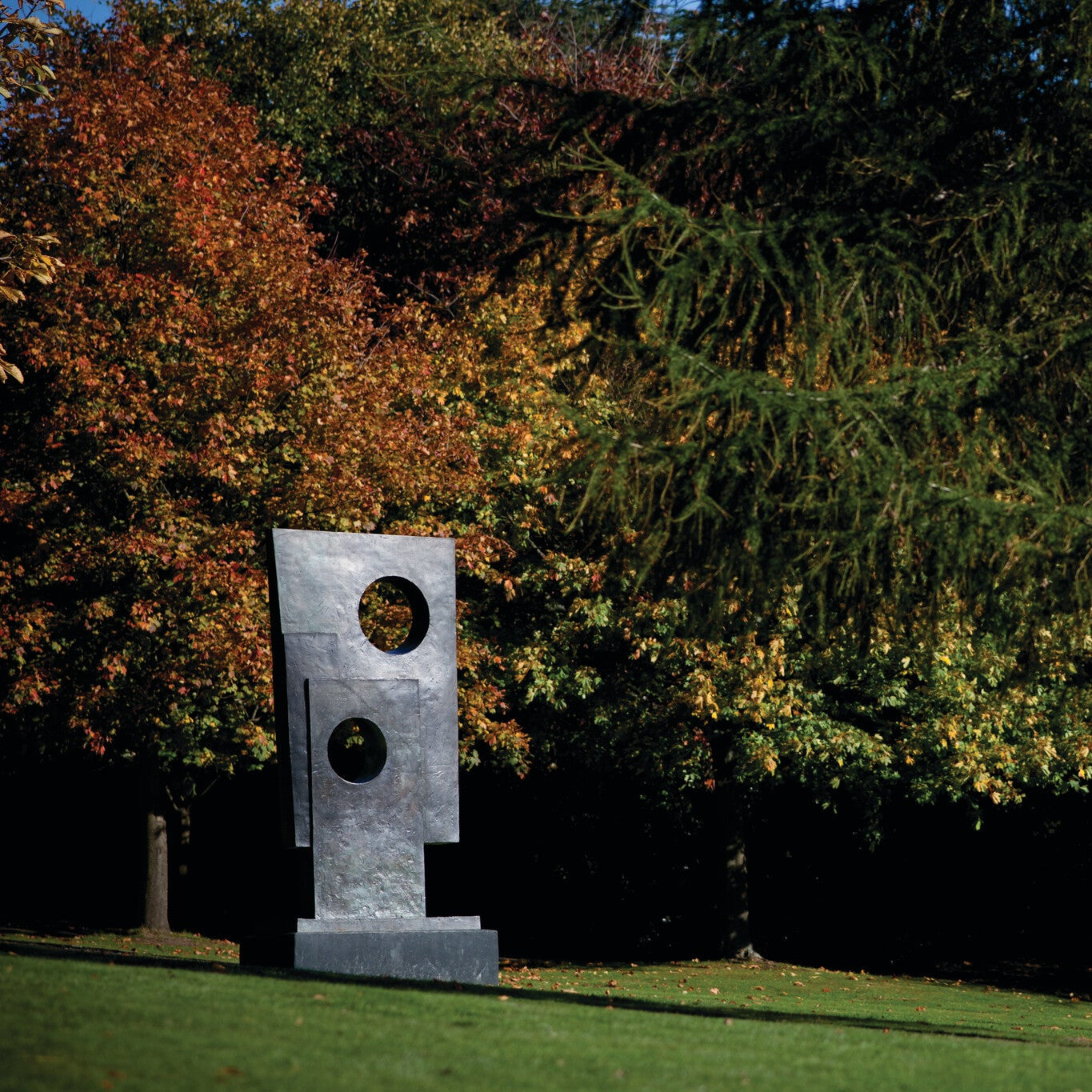 A modern sculpture stands in a park, featuring geometric shapes with circular cutouts. Surrounding it are trees displaying vibrant autumn foliage, creating a picturesque scene of nature and art in harmony.