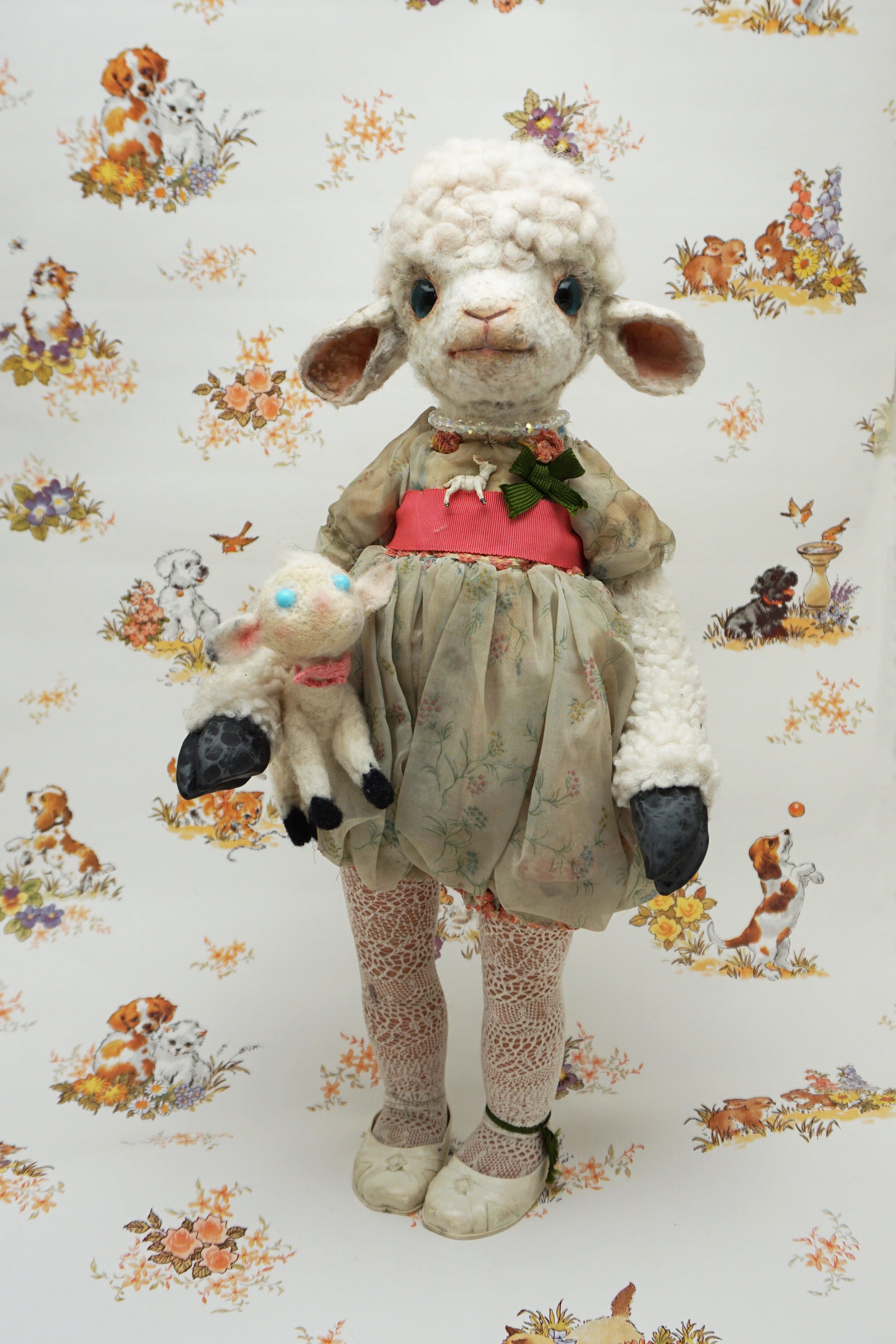 A plush lamb toy dressed in a floral dress with a red sash, holding a smaller lamb toy. The background features a whimsical pattern of animals and flowers, including dogs and rabbits, in soft pastel colours.