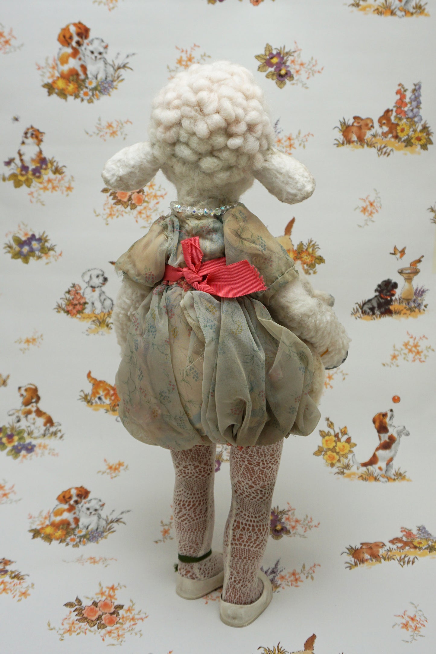 A plush lamb toy dressed in a floral dress with a red sash, holding a smaller lamb toy. The background features a whimsical pattern of animals and flowers, including dogs and rabbits, in soft pastel colours.