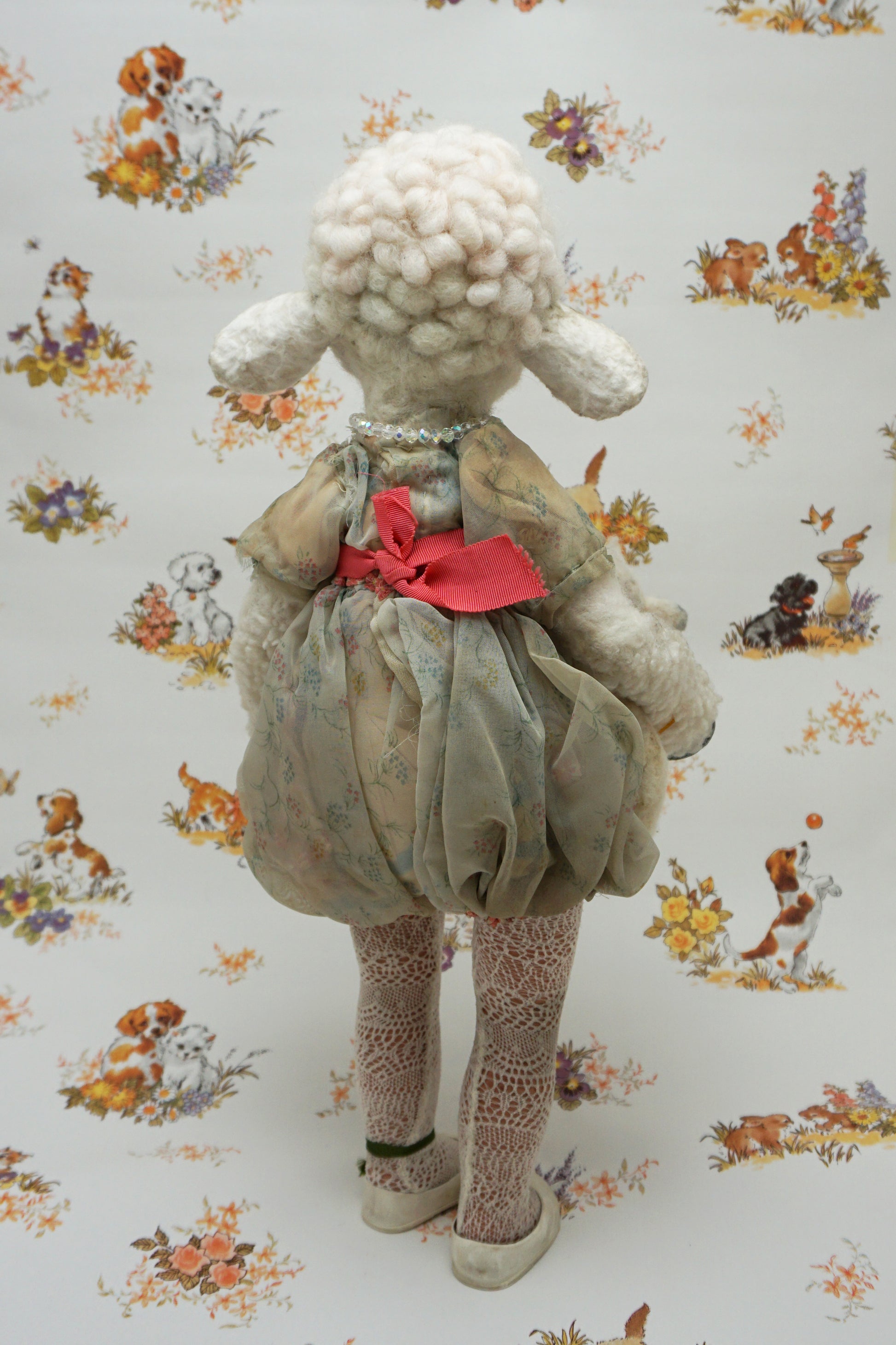 A plush lamb toy dressed in a floral dress with a red sash, holding a smaller lamb toy. The background features a whimsical pattern of animals and flowers, including dogs and rabbits, in soft pastel colours.