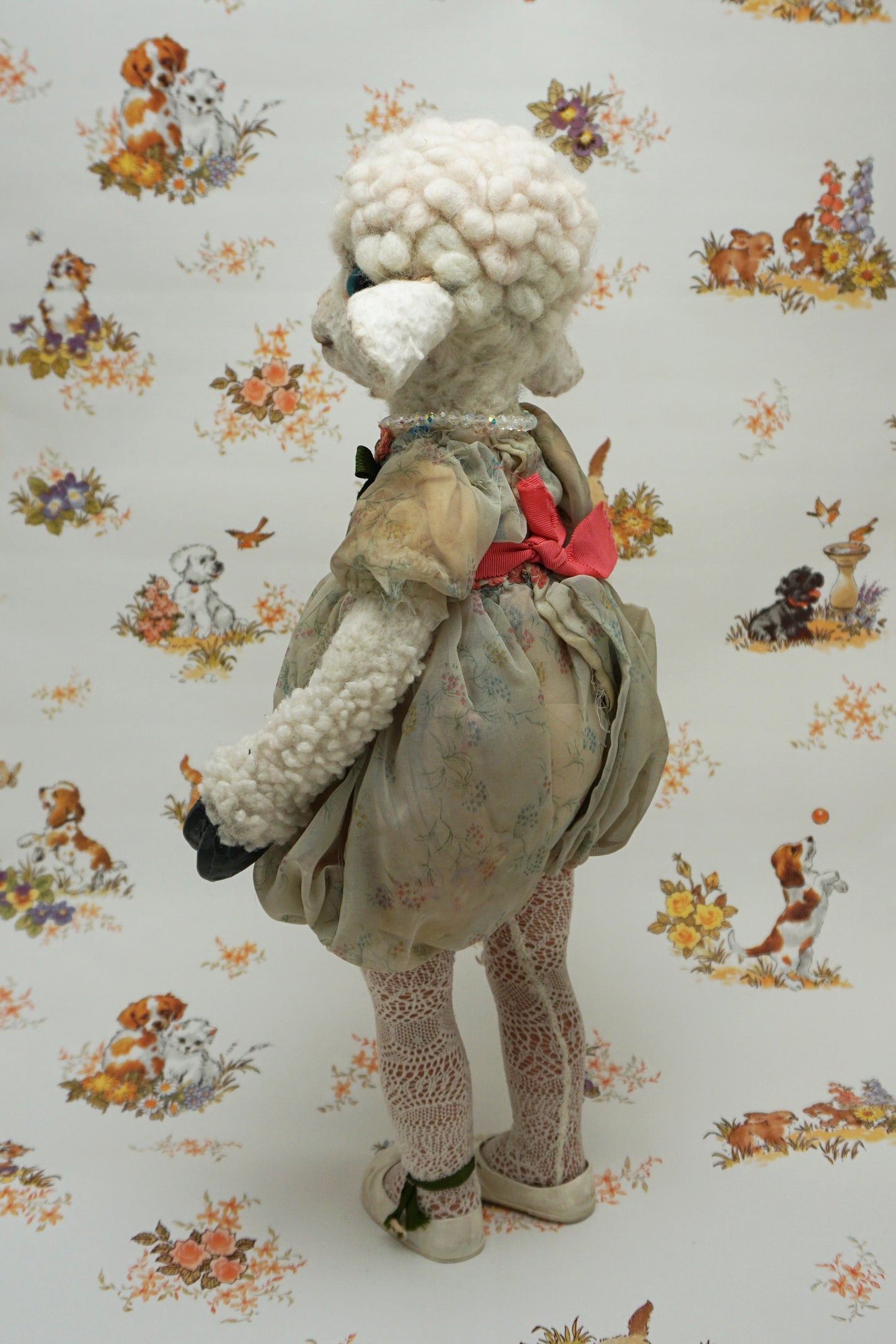A plush lamb toy dressed in a floral dress with a red sash, holding a smaller lamb toy. The background features a whimsical pattern of animals and flowers, including dogs and rabbits, in soft pastel colours.