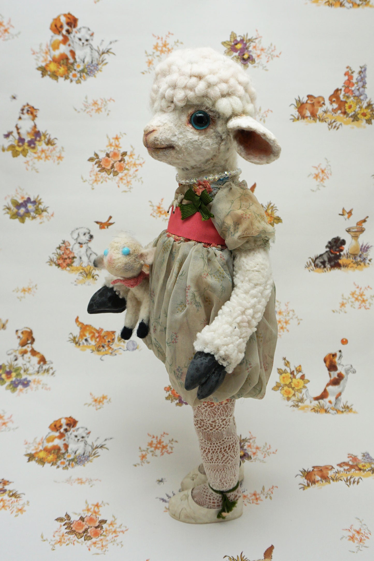 A plush lamb toy dressed in a floral dress with a red sash, holding a smaller lamb toy. The background features a whimsical pattern of animals and flowers, including dogs and rabbits, in soft pastel colours.