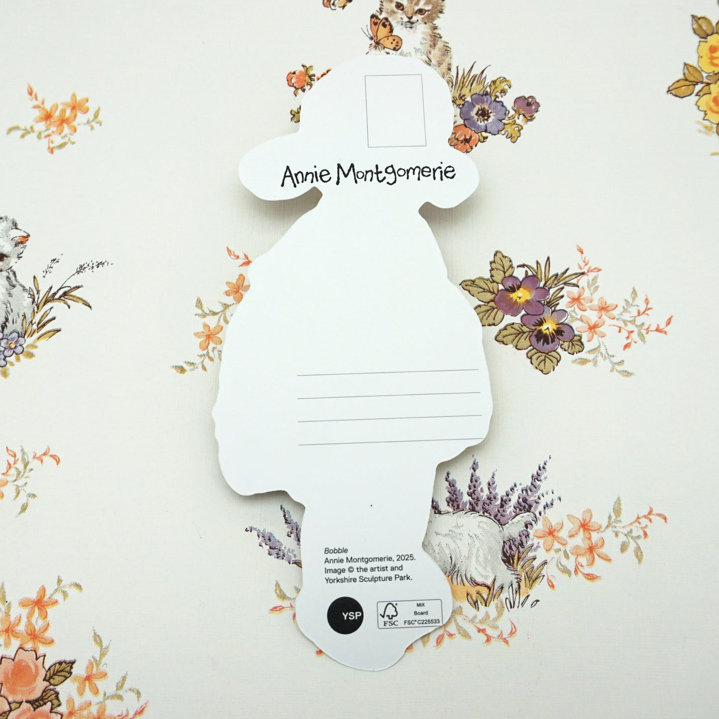 A silhouette-shaped card with "Annie Montgomerie" written on it, set against a floral wallpaper background featuring rabbits and flowers. The card has lines for writing and a small logo at the bottom.