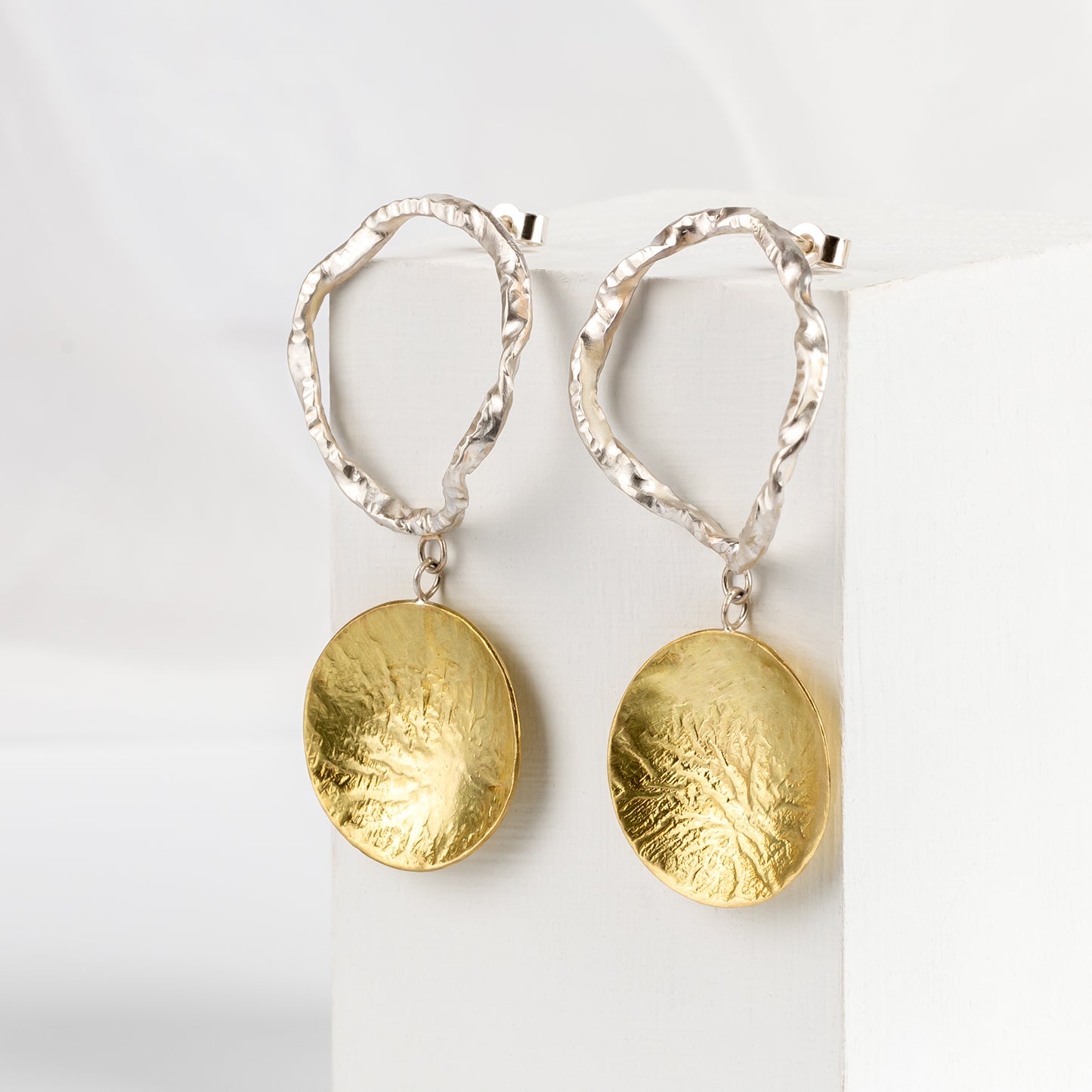A pair of earrings featuring a silver circular drop and brass disc design, displayed against a white background.
