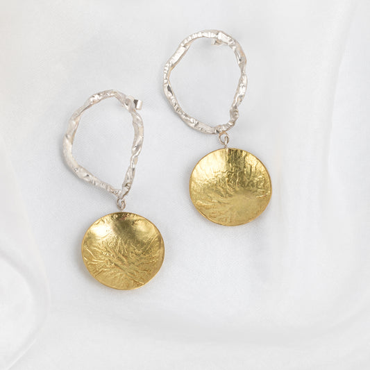 A pair of earrings featuring a silver circular drop and brass disc design, displayed against a white background.