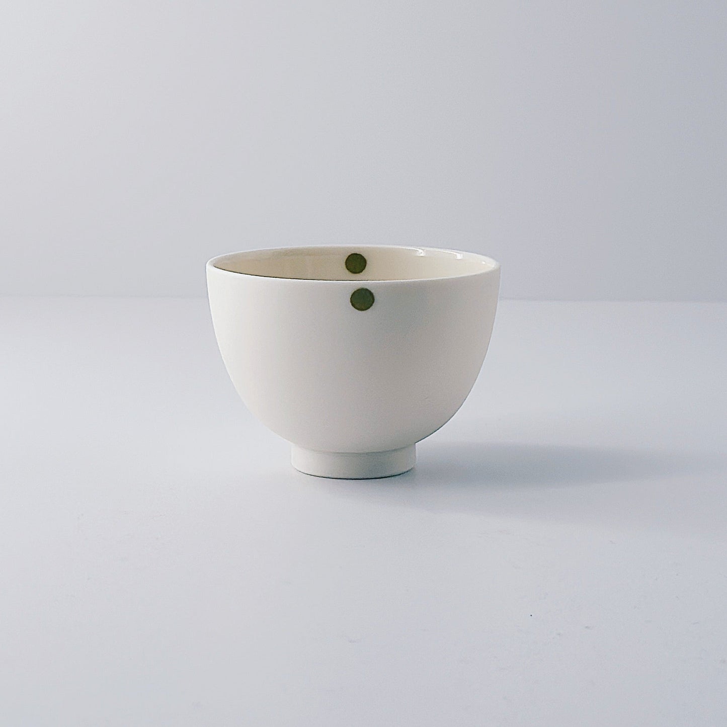 White porcelain bowl with a single olive green dot on a light grey background