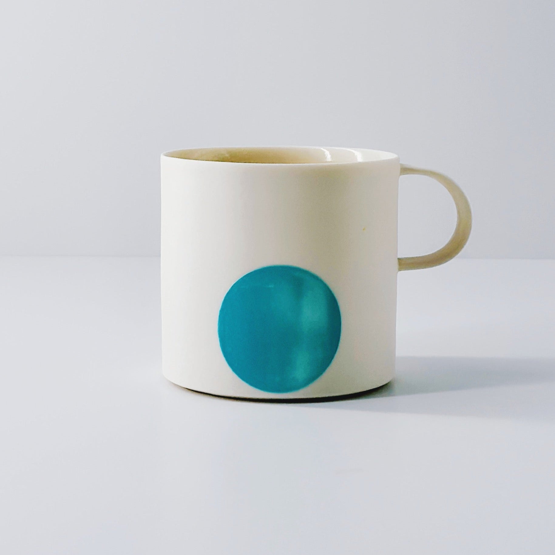 White porcelain mug with a blue circle and a dainty handle on a light grey background.