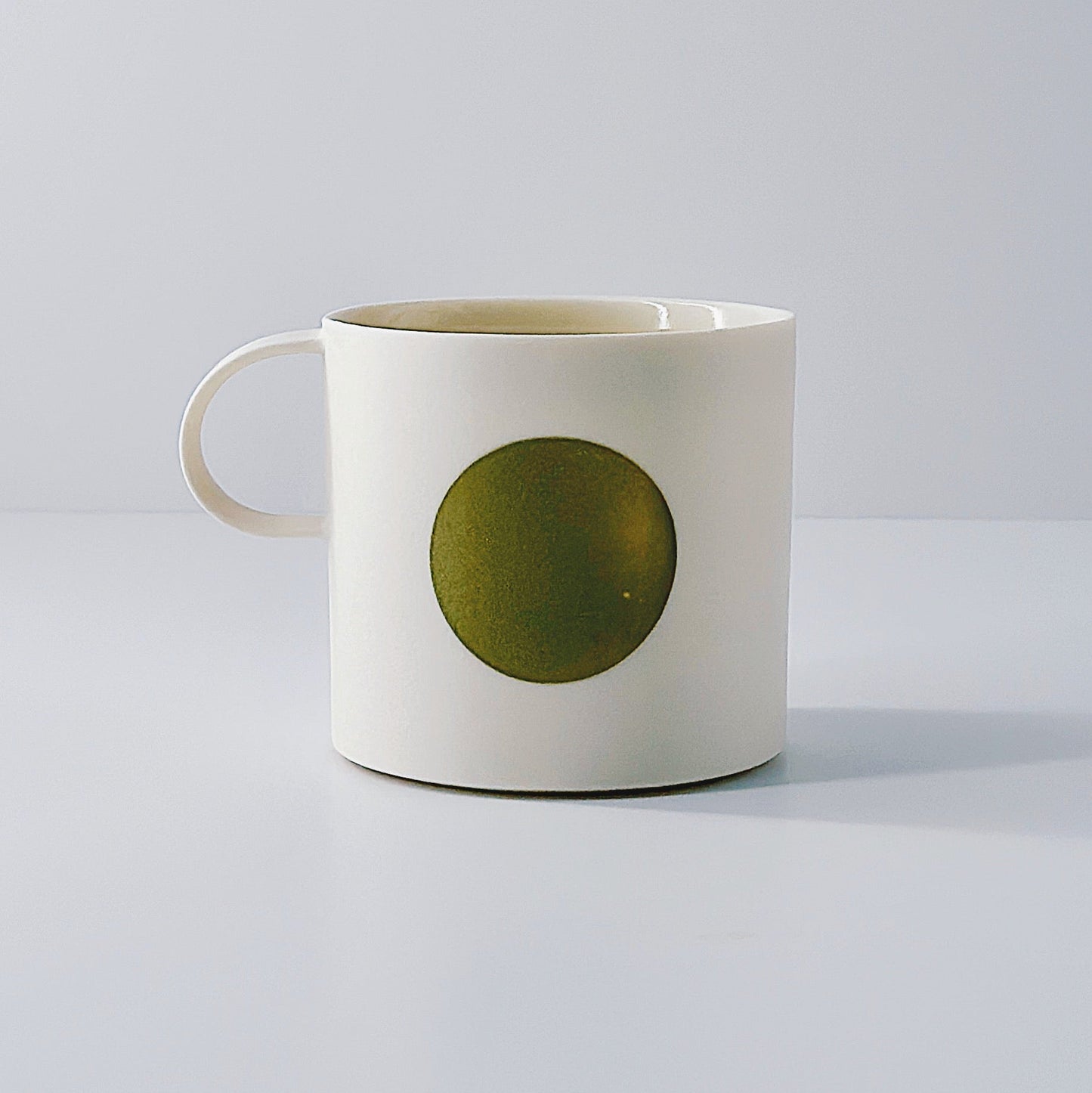 White porcelain mug with an olive green circle and a dainty handle on a light grey background.