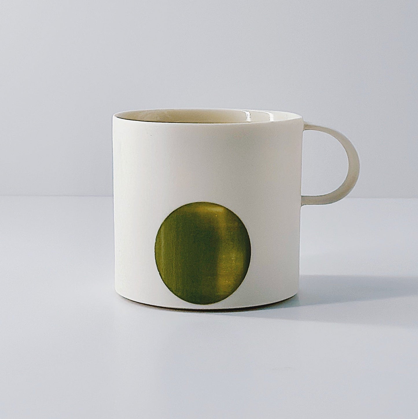 White porcelain mug with an olive green circle and a dainty handle on a light grey background.