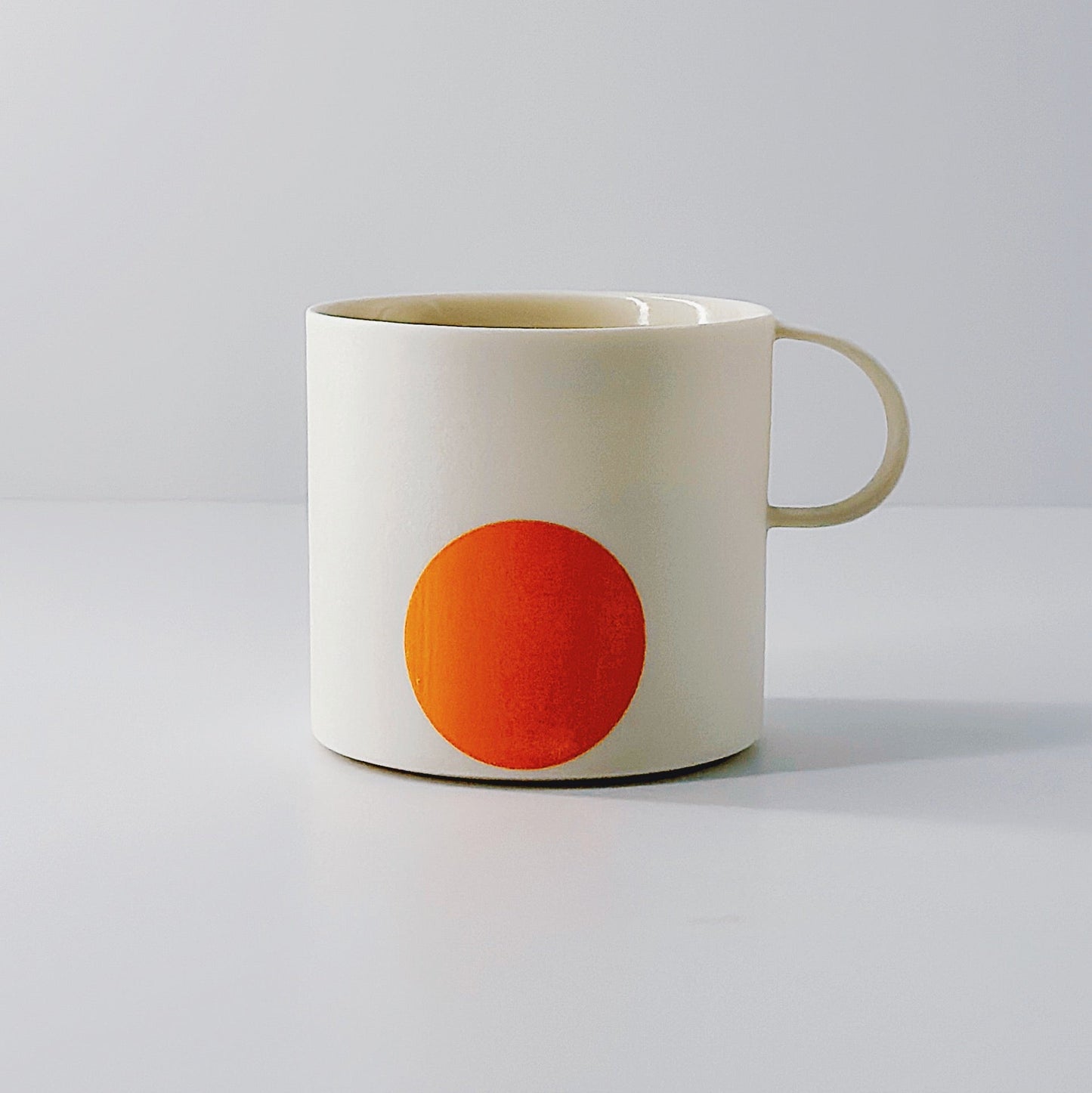 White porcelain mug with an orange circle and a dainty handle on a light grey background.