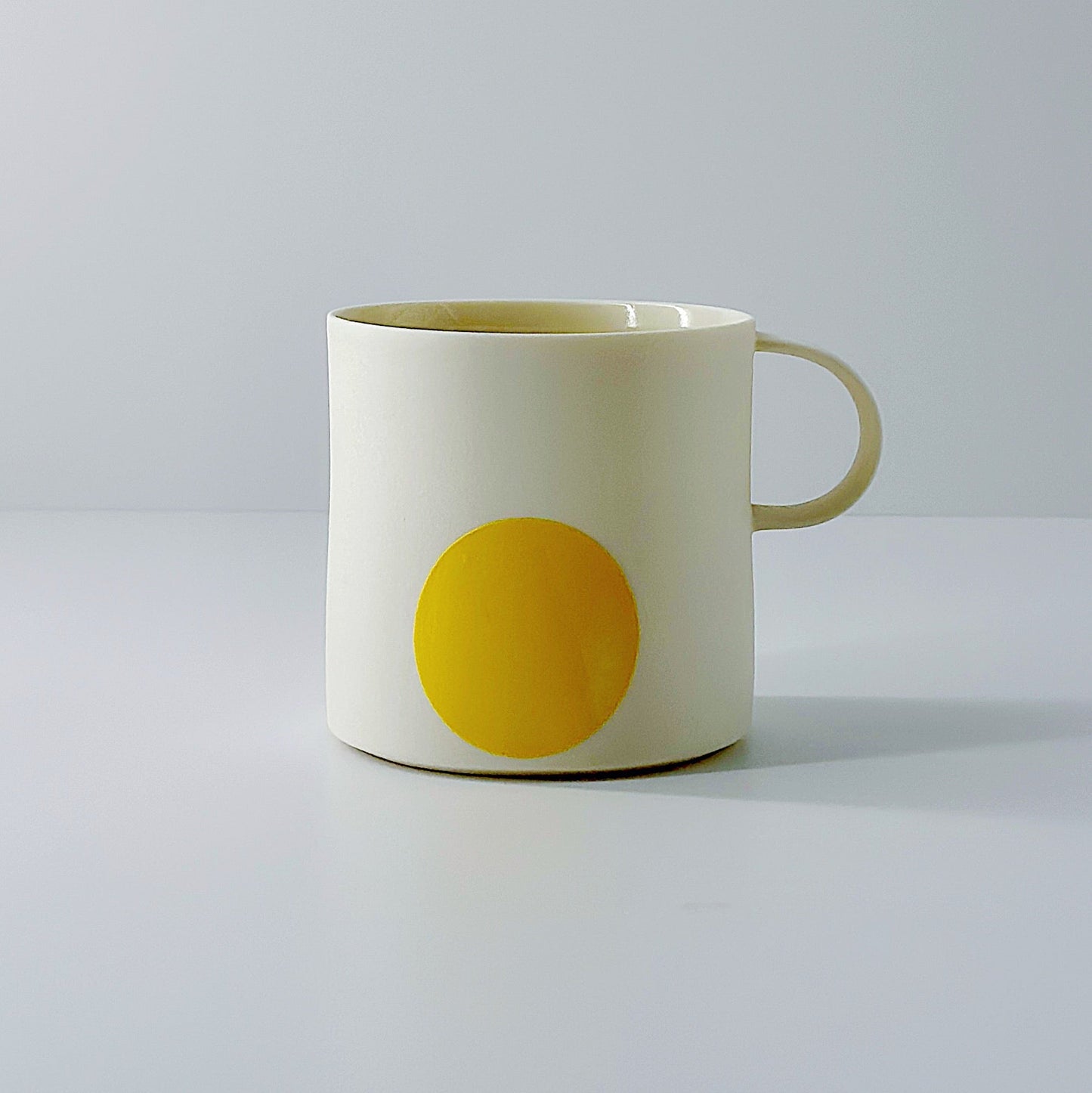 White porcelain mug with a yellow circle and a dainty handle on a light grey background.