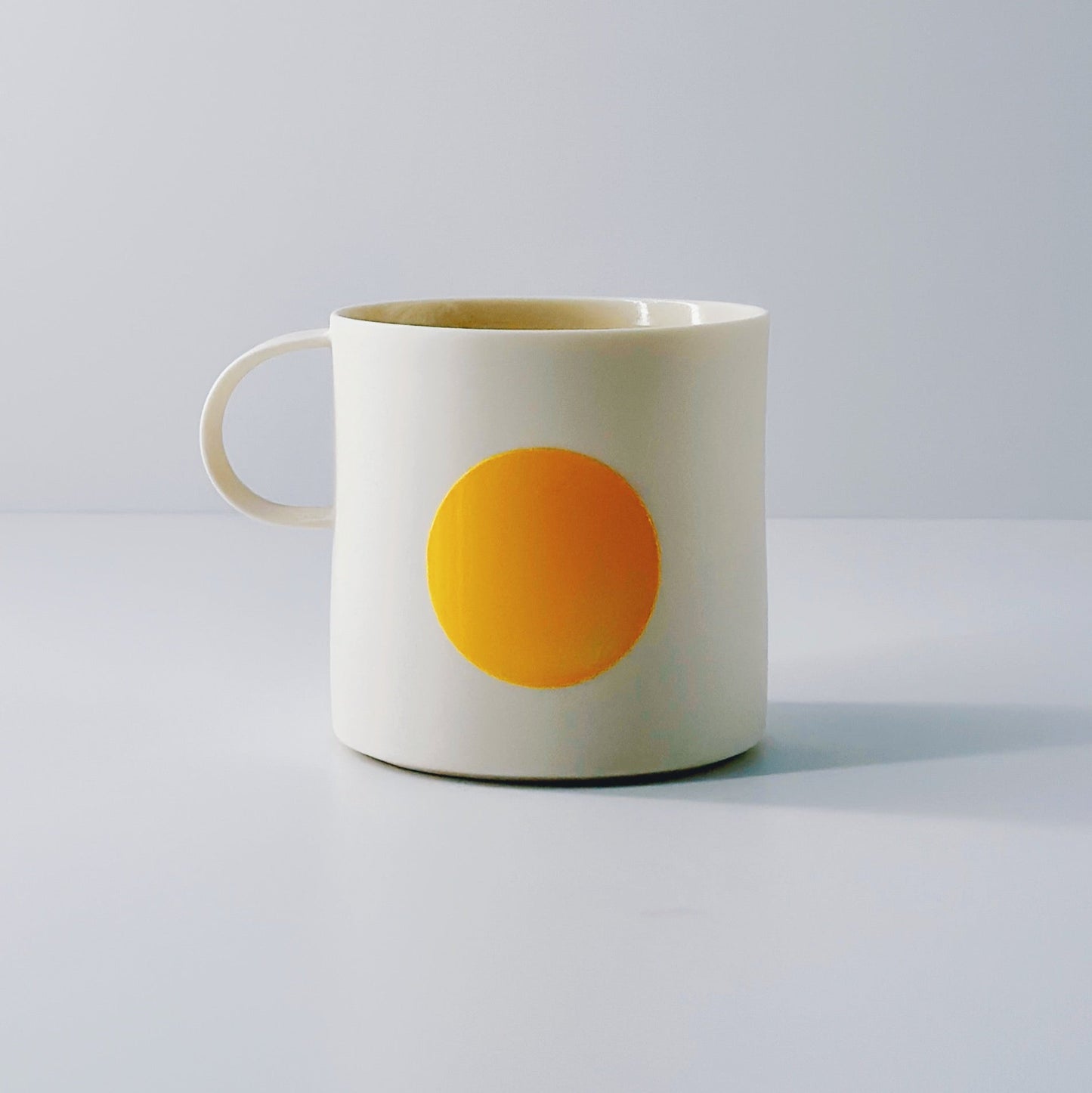 White porcelain mug with a yellow circle and a dainty handle on a light grey background.