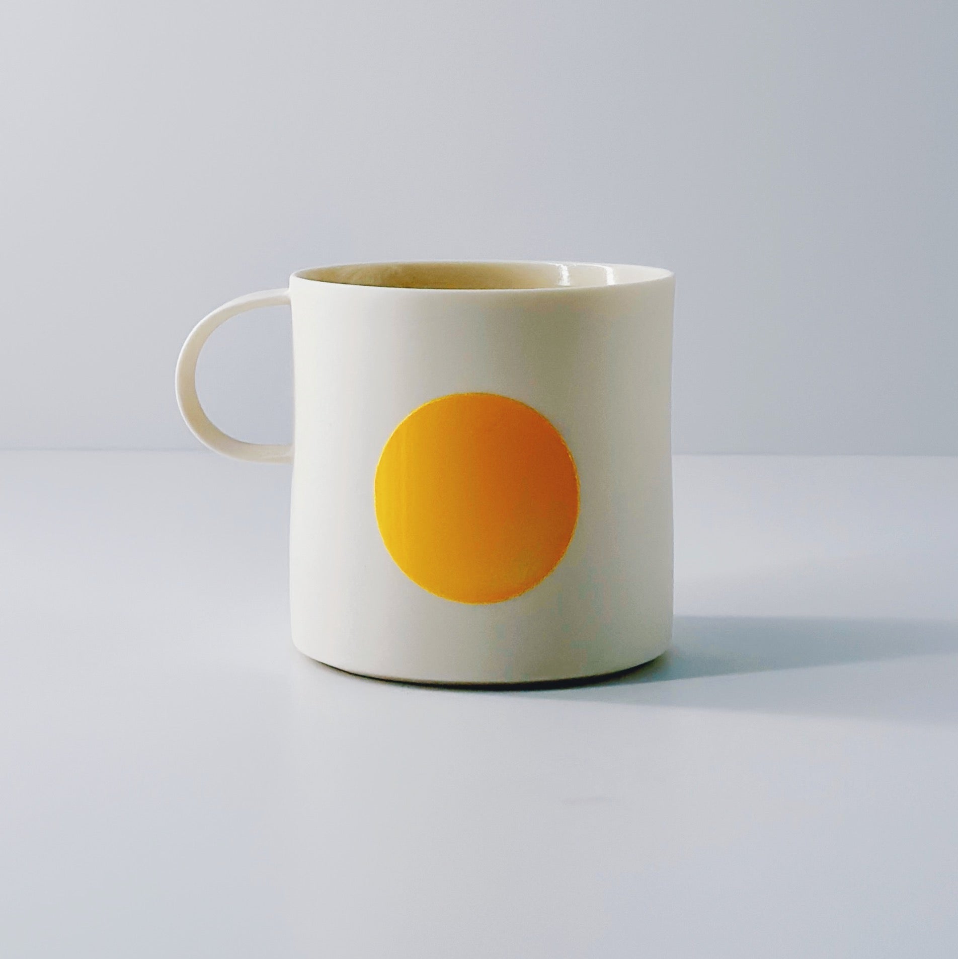 White porcelain mug with a yellow circle and a dainty handle on a light grey background.