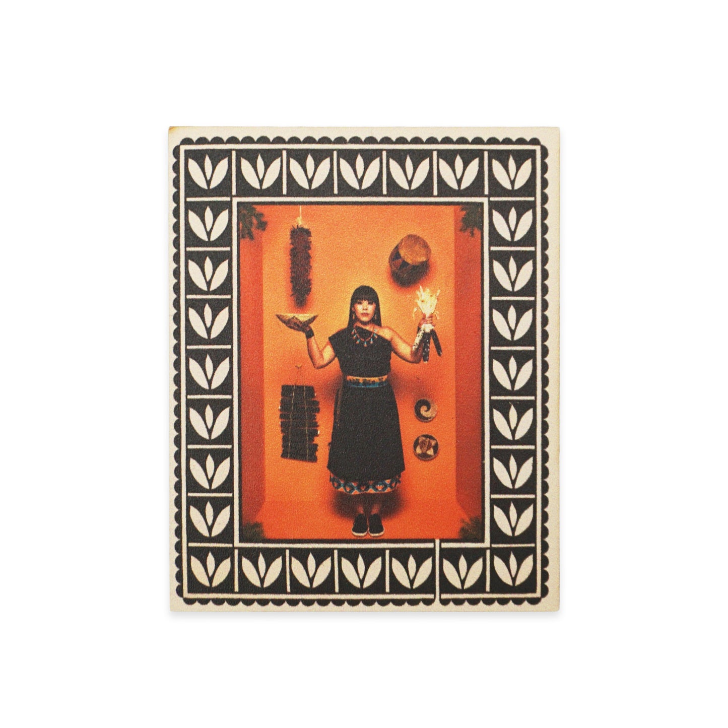 A woman stands against an orange background, holding a bird and a doll. She wears a black dress with decorative trim. The image is framed with a black and white leaf pattern. Various objects hang on the wall, including a hat and a crescent moon.