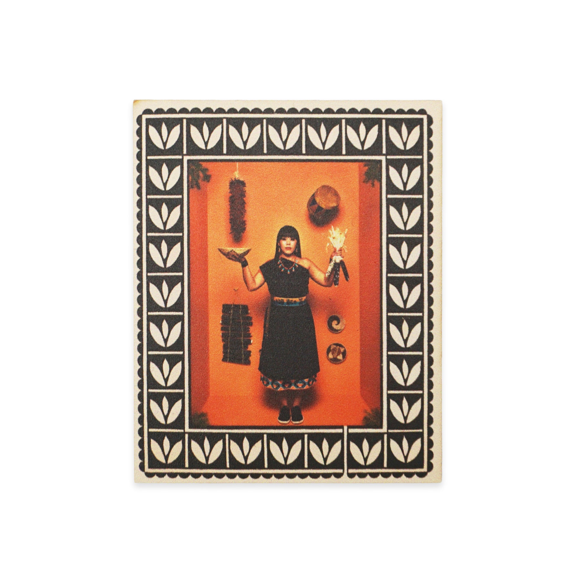 A woman stands against an orange background, holding a bird and a doll. She wears a black dress with decorative trim. The image is framed with a black and white leaf pattern. Various objects hang on the wall, including a hat and a crescent moon.
