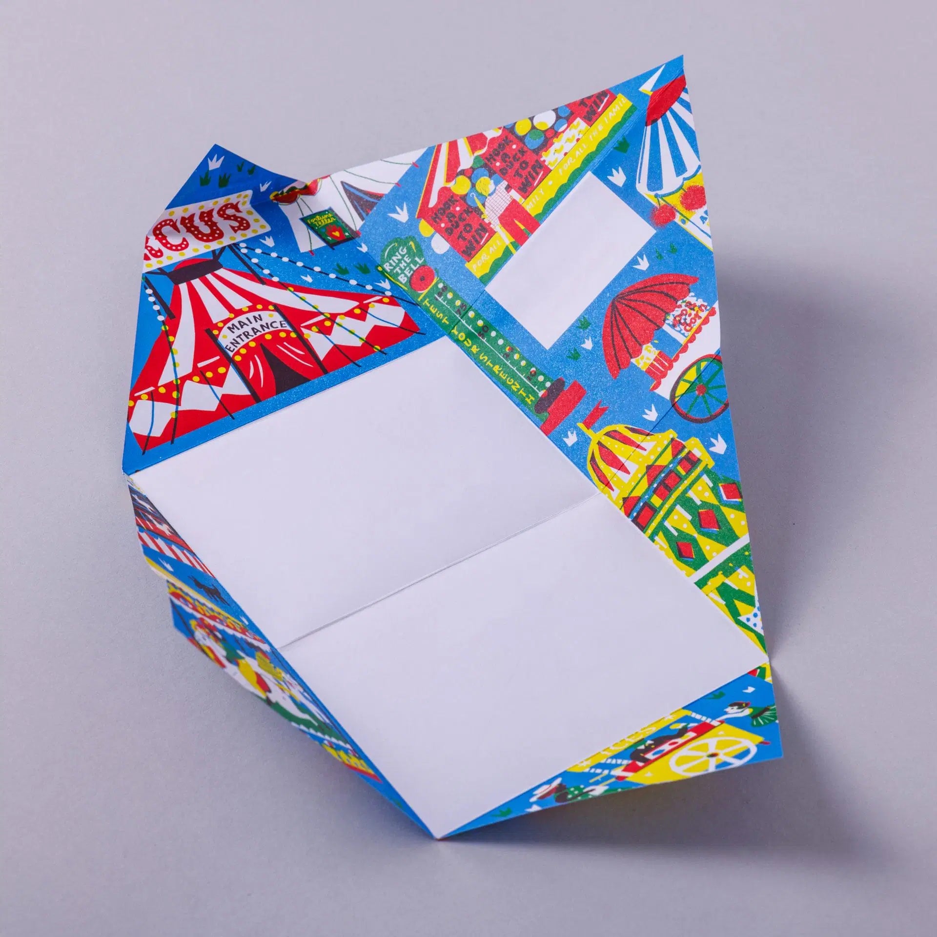 A colourful origami paper featuring a vibrant circus theme with tents, carousels, and balloons. The inside is blank, ready for a message, while the exterior showcases playful illustrations in bright colours.