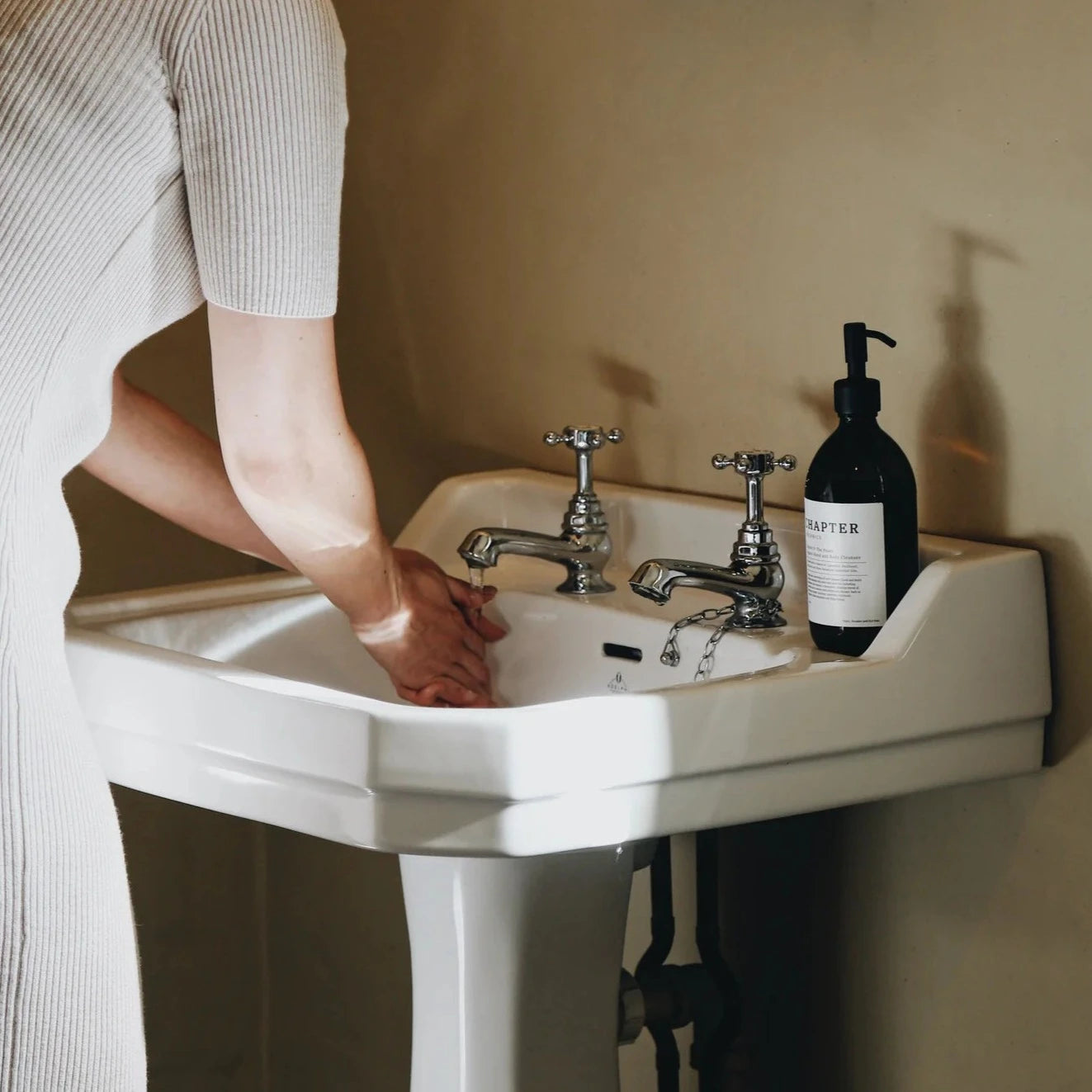 A woman in a fitted white dress washes her hands at a vintage-style sink. The sink features two taps and a soap dispenser beside it. A mirror hangs on the wall, reflecting the scene in a softly lit, neutral-toned room.