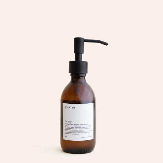 A brown glass pump bottle with a black pump lid. The label features minimalist text, indicating the product name "The Quiet" and a brief description. The background is a soft, neutral colour, enhancing the bottle's elegant design.