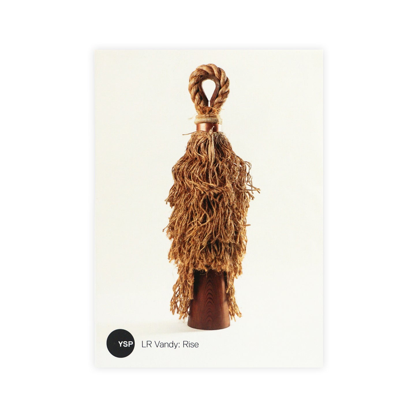 A decorative object featuring a wooden base with a twisted rope handle. The top is adorned with long, textured strands resembling a tassel, creating a rustic and artistic appearance. The background is plain and light-coloured.