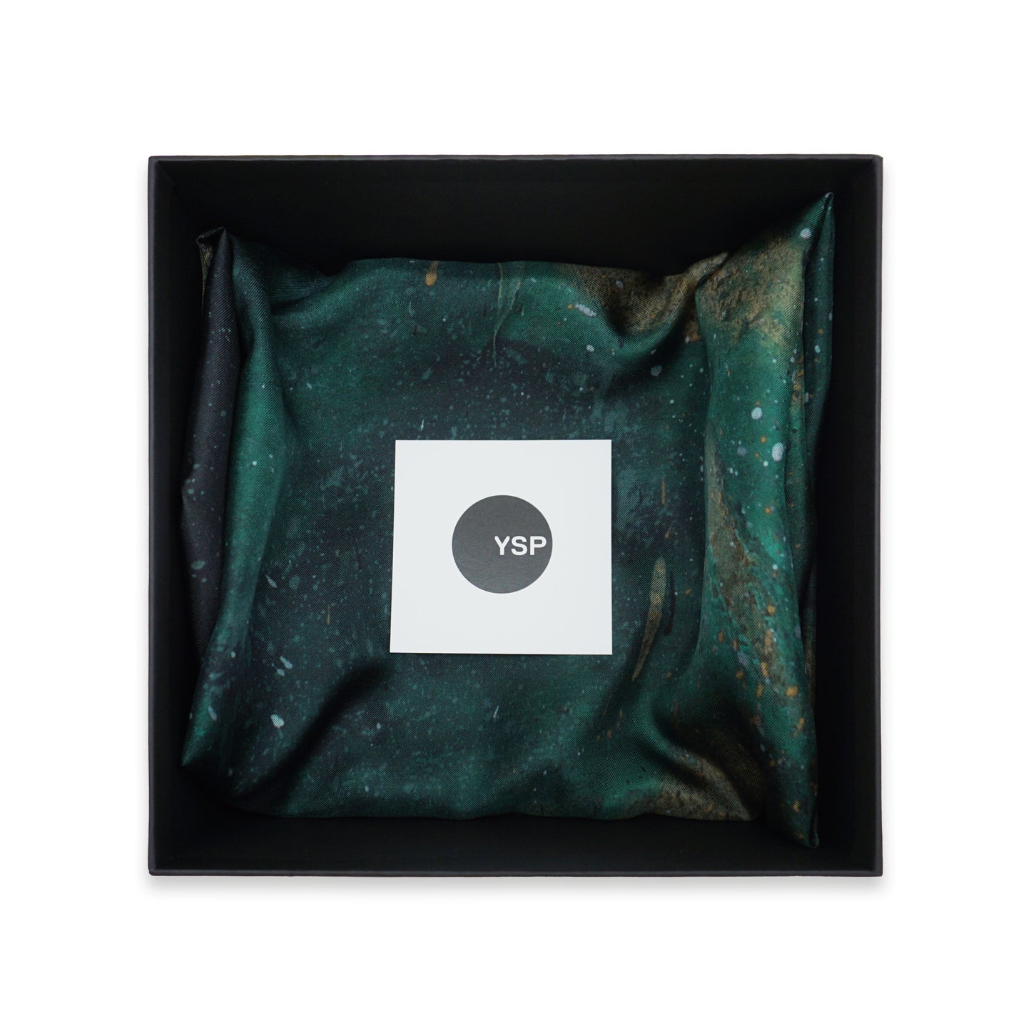 A square black gift box containing a folded dark green and black patterned fabric item with a white square label in the centre that reads 'YSP'.