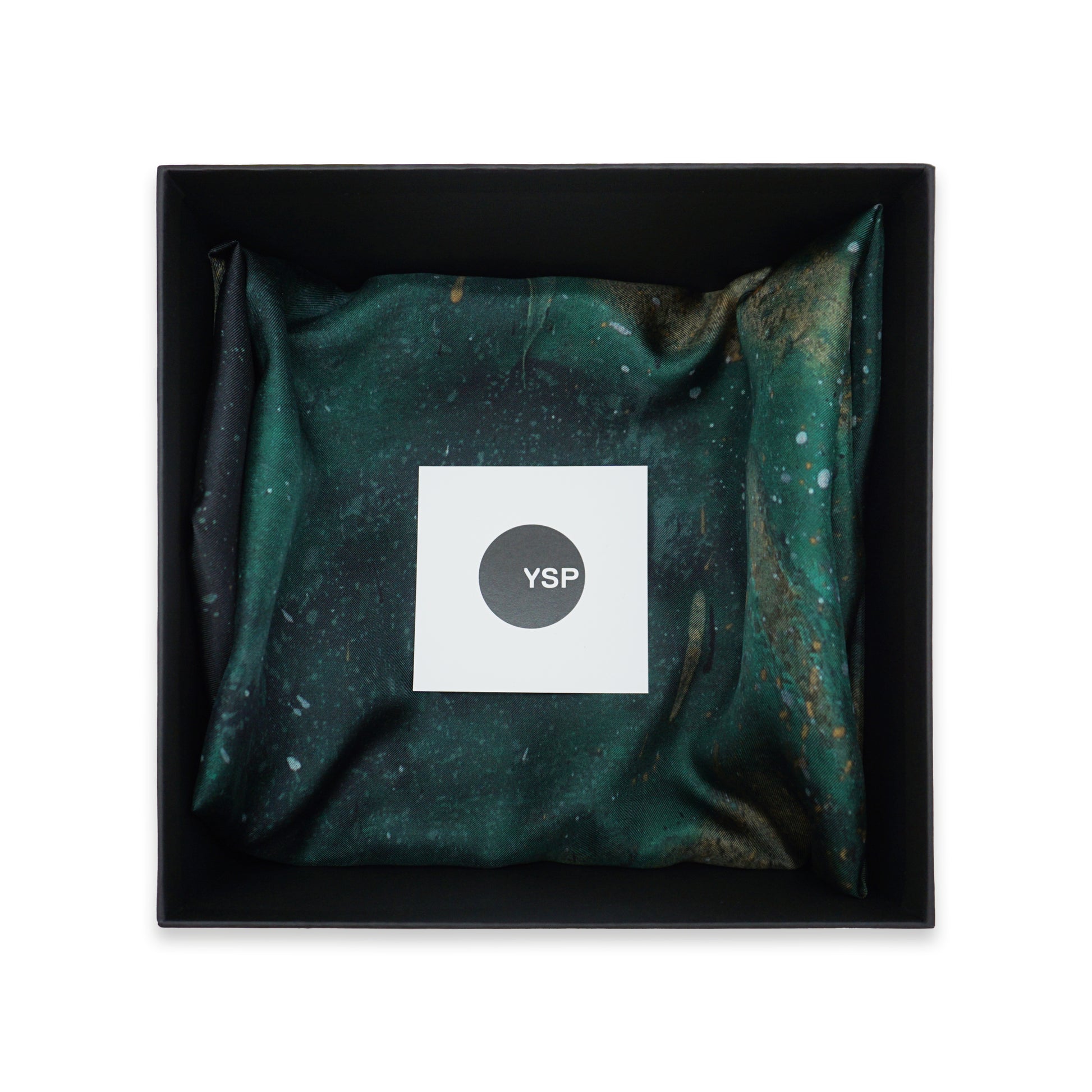 A square black gift box containing a folded dark green and black patterned fabric item with a white square label in the centre that reads 'YSP'.