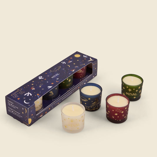 A set of four small candles with a matching dark blue outer box decorated with gold celestial illustrations. The candles are in different colored glass containers: cream, dark blue, green, and burgundy, all featuring gold star and constellation patterns.