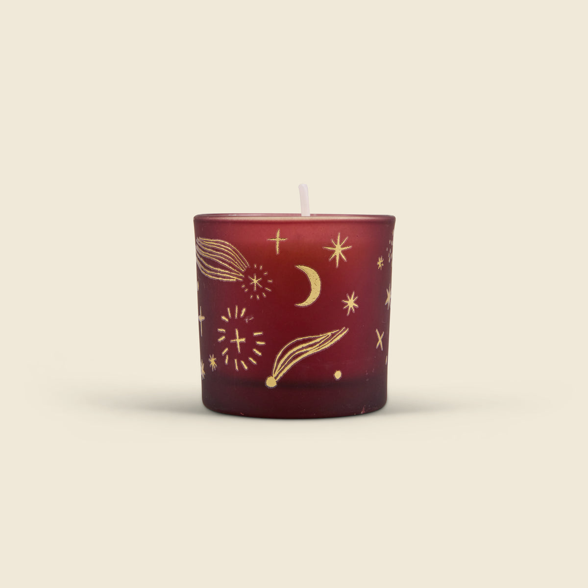 A small red glass candle with a white wick, decorated with gold celestial illustrations including stars, comets, and a crescent moon, set against a solid light beige background.