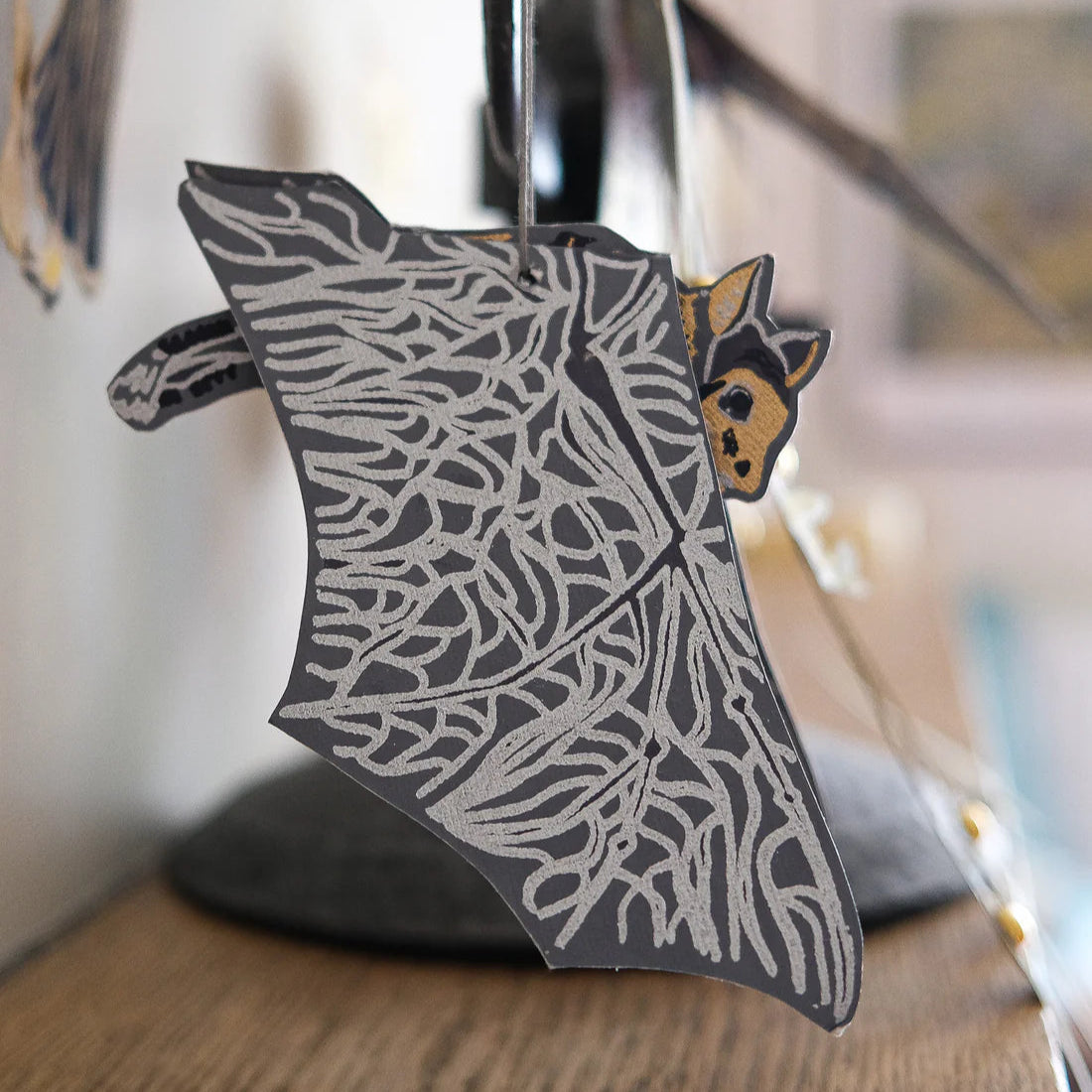 Decorative bat in flight paper cut-out with intricate patterns on a wooden surface. The main colours are grey and beige.