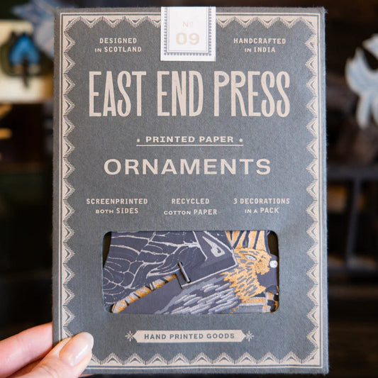 Packaging of East End Press bat ornaments held by a hand with a blurred background. The packaging is grey with beige text.