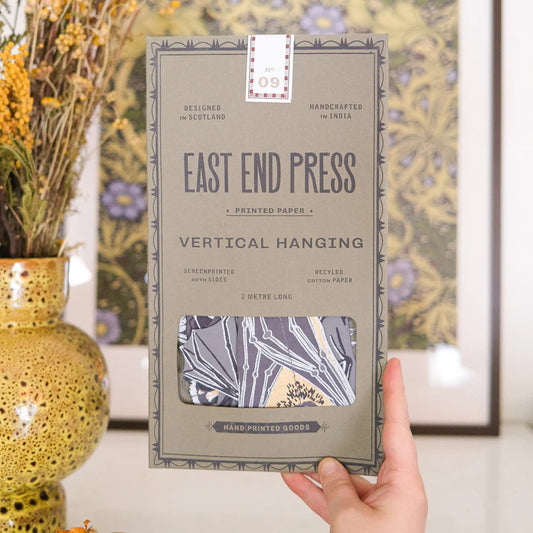 Handmade paper packaging with 'East End Press' branding held by a hand, with decorative elements in the background. The packaging is grey with blue text.