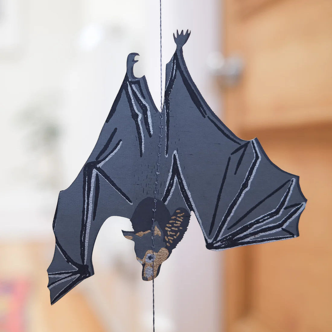 Decorative paper bat hanging on a string with a blurred background. The bat is grey with blue and beige accents.