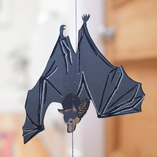 Decorative paper bat hanging on a string with a blurred background. The bat is grey with blue and beige accents.