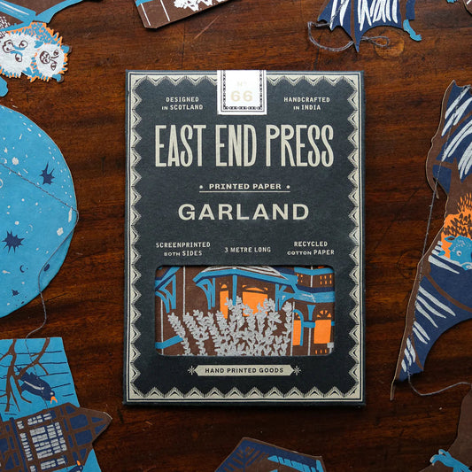East End Press Garland printed paper packaging on a wooden surface with decorative paper cut-outs. The cut-outs feature bats, moons and houses all in shades of blue, brown and orange.