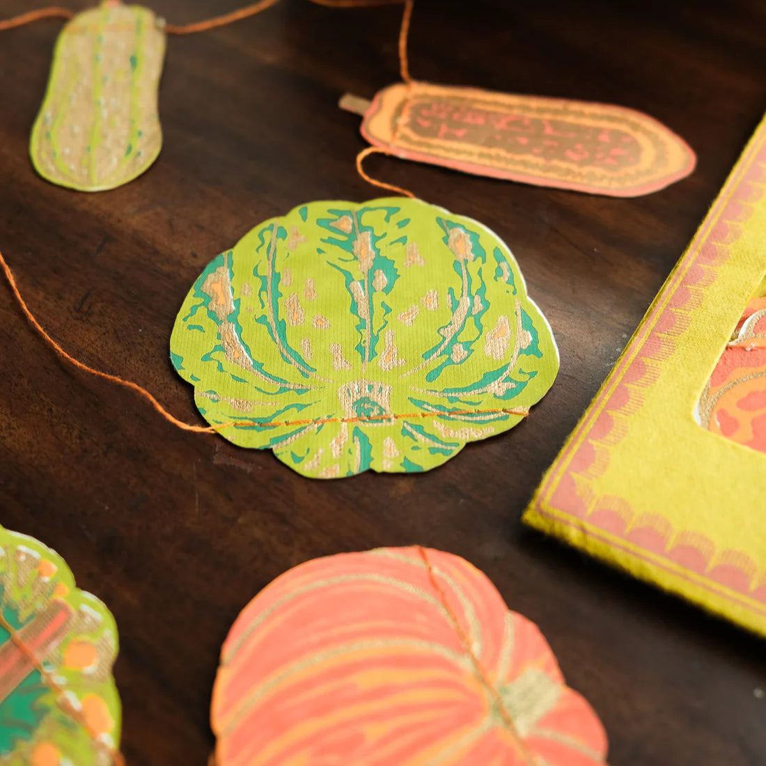 Colourful paper orange and green pumpkins and gourds cut-outs on a wooden surface with a pumpkin in the background.