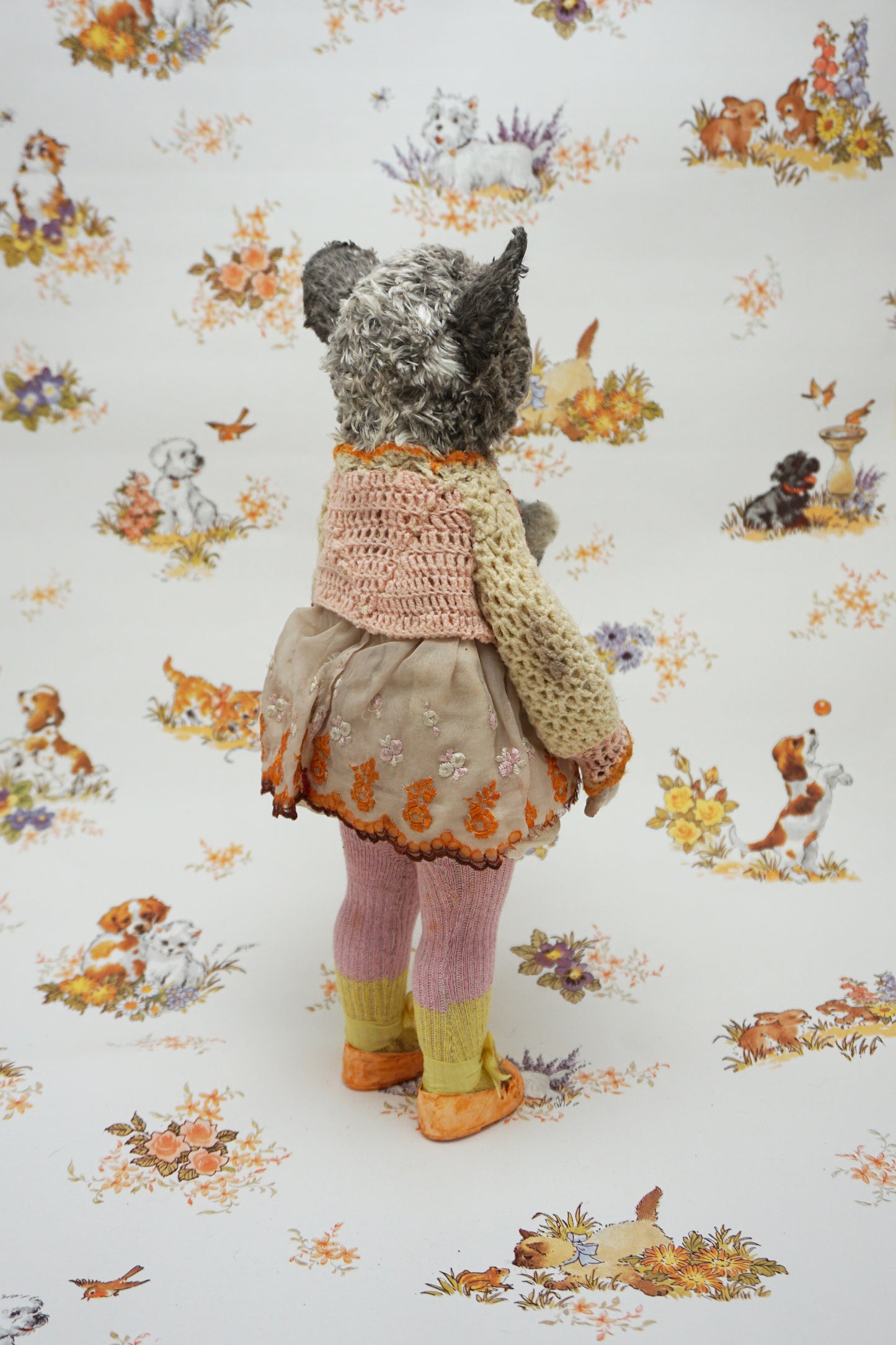A plush toy resembling a bush baby stands against a patterned background. It wears a knitted dress and holds a small bear toy. The backdrop features whimsical animal illustrations in soft colours.