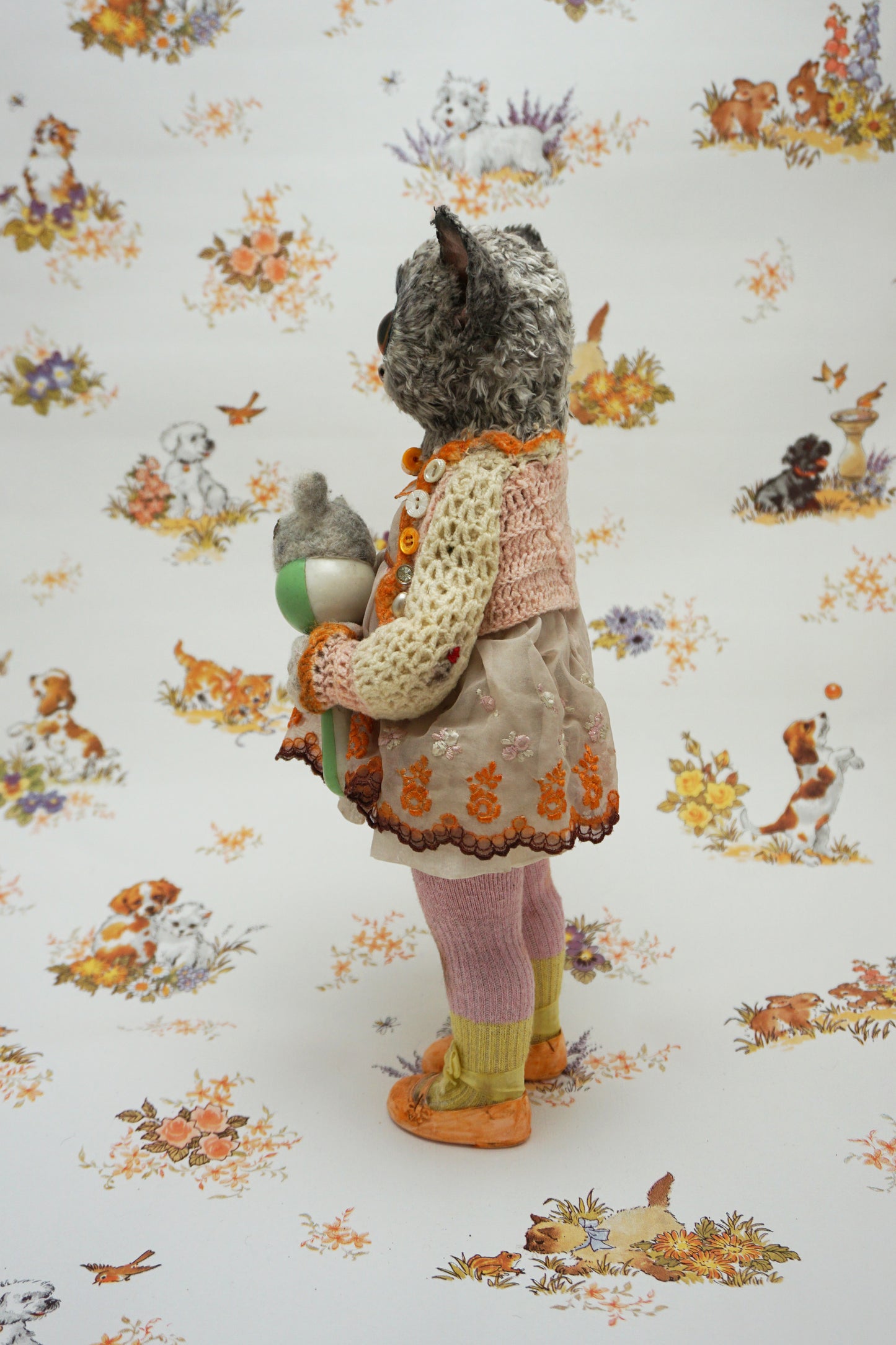 A plush toy resembling a bush baby stands against a patterned background. It wears a knitted dress and holds a small bear toy. The backdrop features whimsical animal illustrations in soft colours.