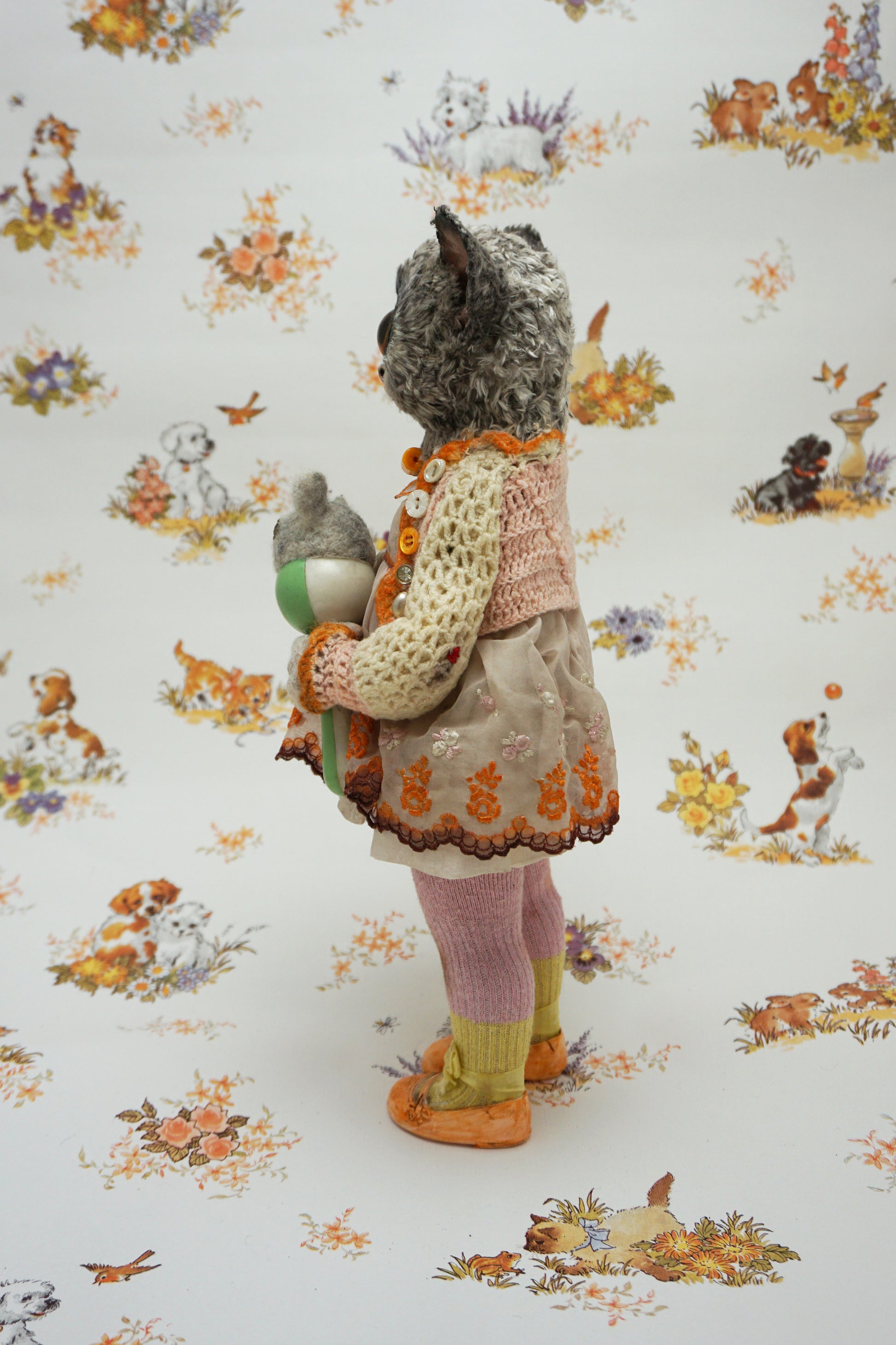 A plush toy resembling a bush baby stands against a patterned background. It wears a knitted dress and holds a small bear toy. The backdrop features whimsical animal illustrations in soft colours.