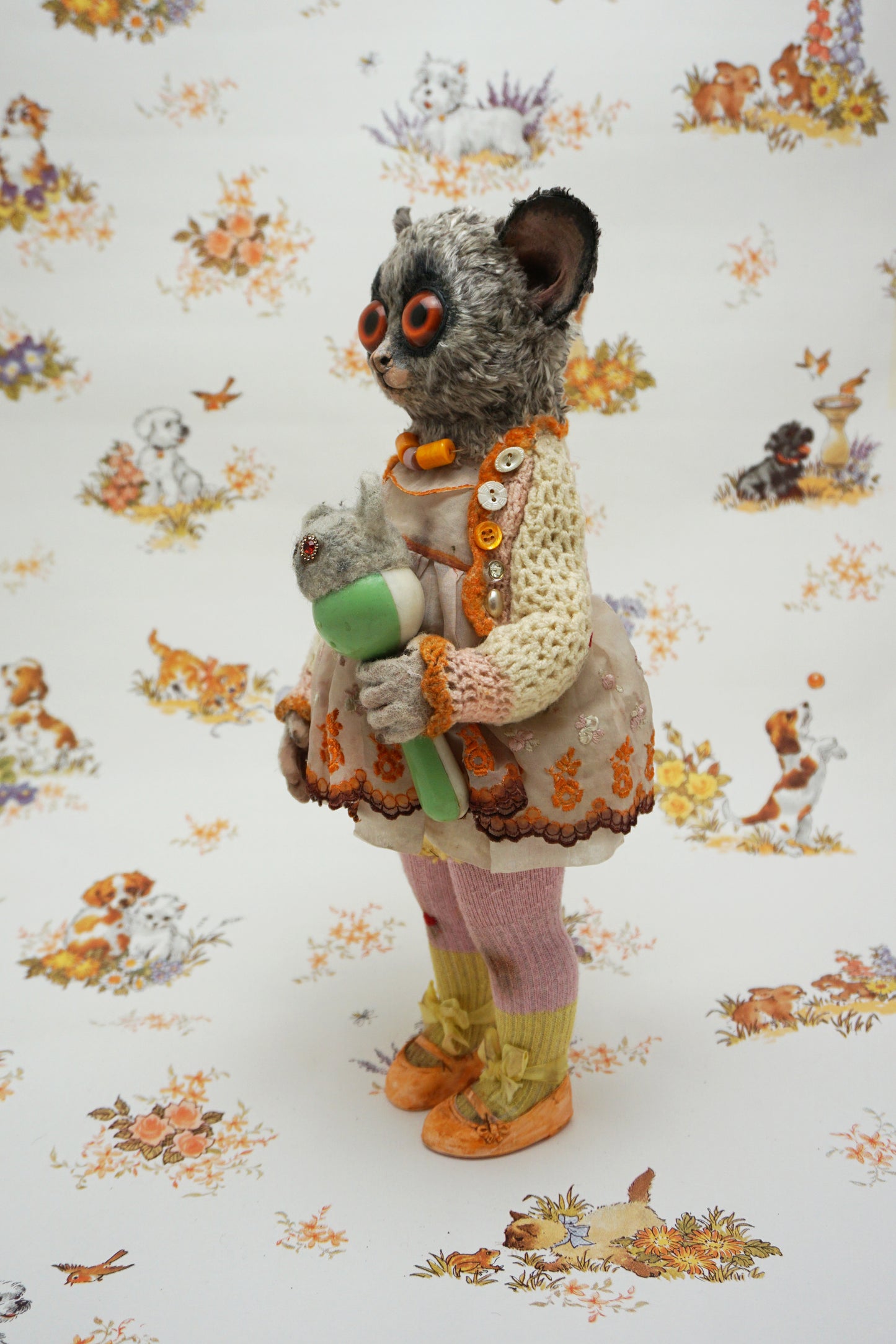 A plush toy resembling a bush baby stands against a patterned background. It wears a knitted dress and holds a small bear toy. The backdrop features whimsical animal illustrations in soft colours.