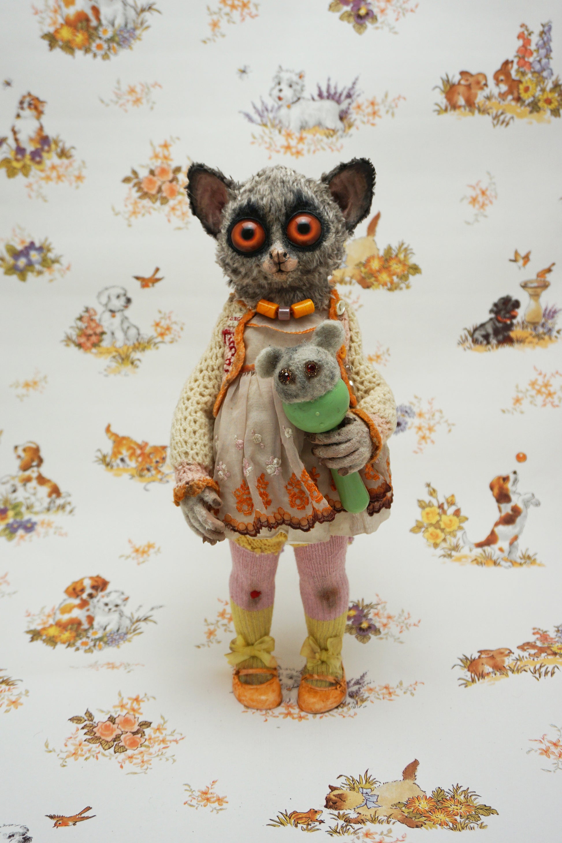 A plush toy resembling a bush baby stands against a patterned background. It wears a knitted dress and holds a small bear toy. The backdrop features whimsical animal illustrations in soft colours.