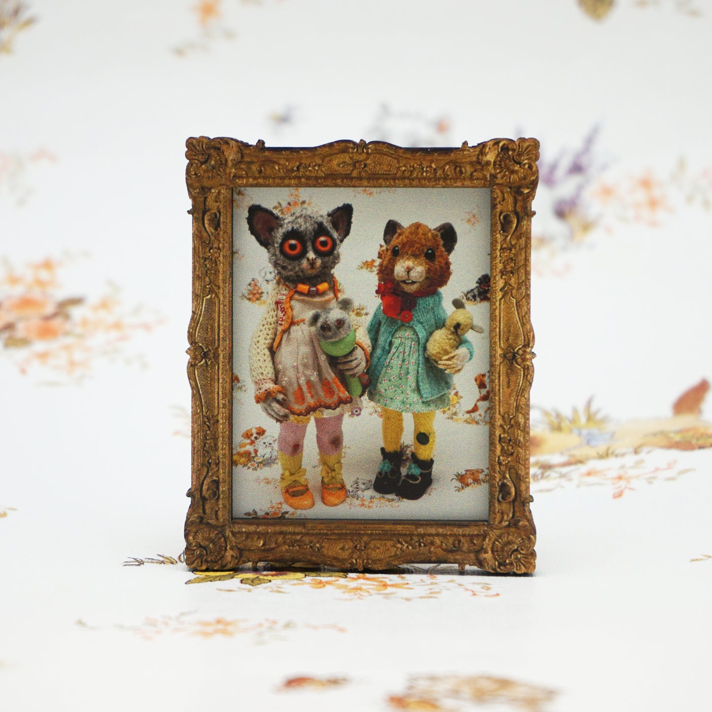 A small, ornate gold frame displays an illustration of two whimsical animal characters dressed in vintage-style clothing. The background features a soft floral pattern, complementing the charming and nostalgic theme of the artwork.