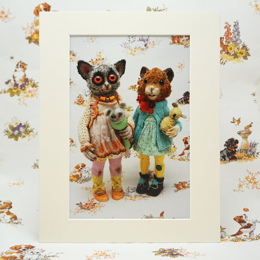 A mini print showing two whimsical animals standing side by side, dressed in colourful, vintage-style clothing. One holds a small teddy bear, while the other carries a tiny doll. The background features a matching pattern of playful animal illustrations. The print has a cream coloured mount to it.