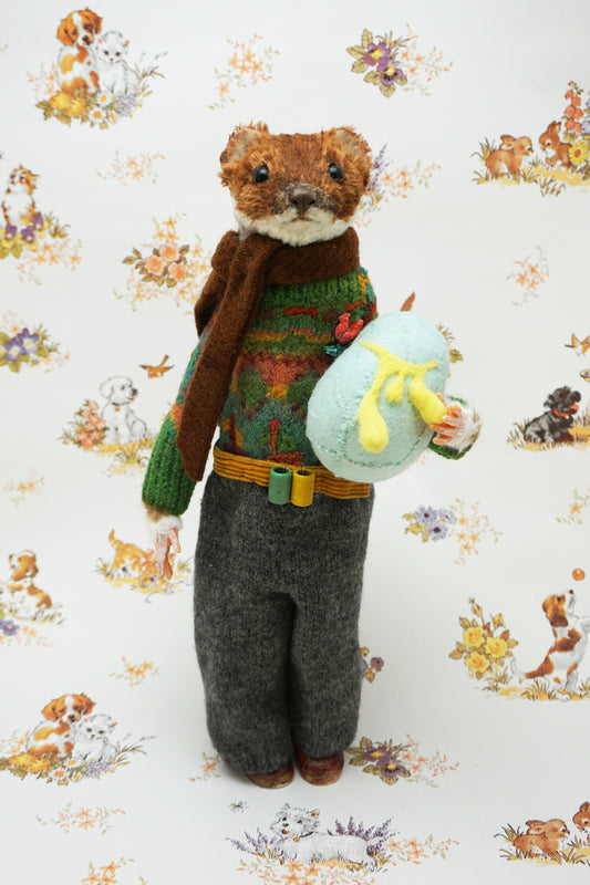 A plush weasel stands upright, wearing a green jumper, brown scarf, and grey trousers. It holds a light blue egg with yellow details. The background features a whimsical pattern of small animals and flowers.