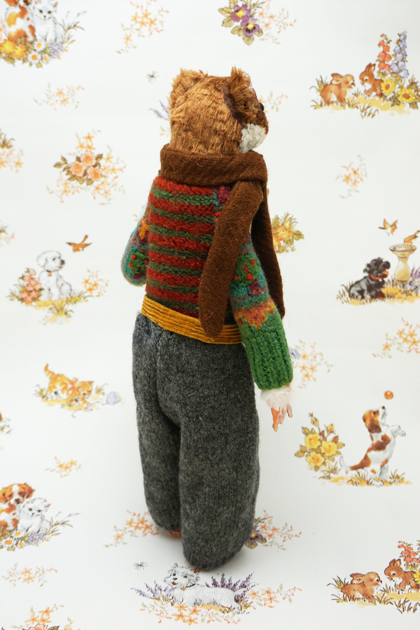A plush weasel stands upright, wearing a green jumper, brown scarf, and grey trousers. It holds a light blue egg with yellow details. The background features a whimsical pattern of small animals and flowers.