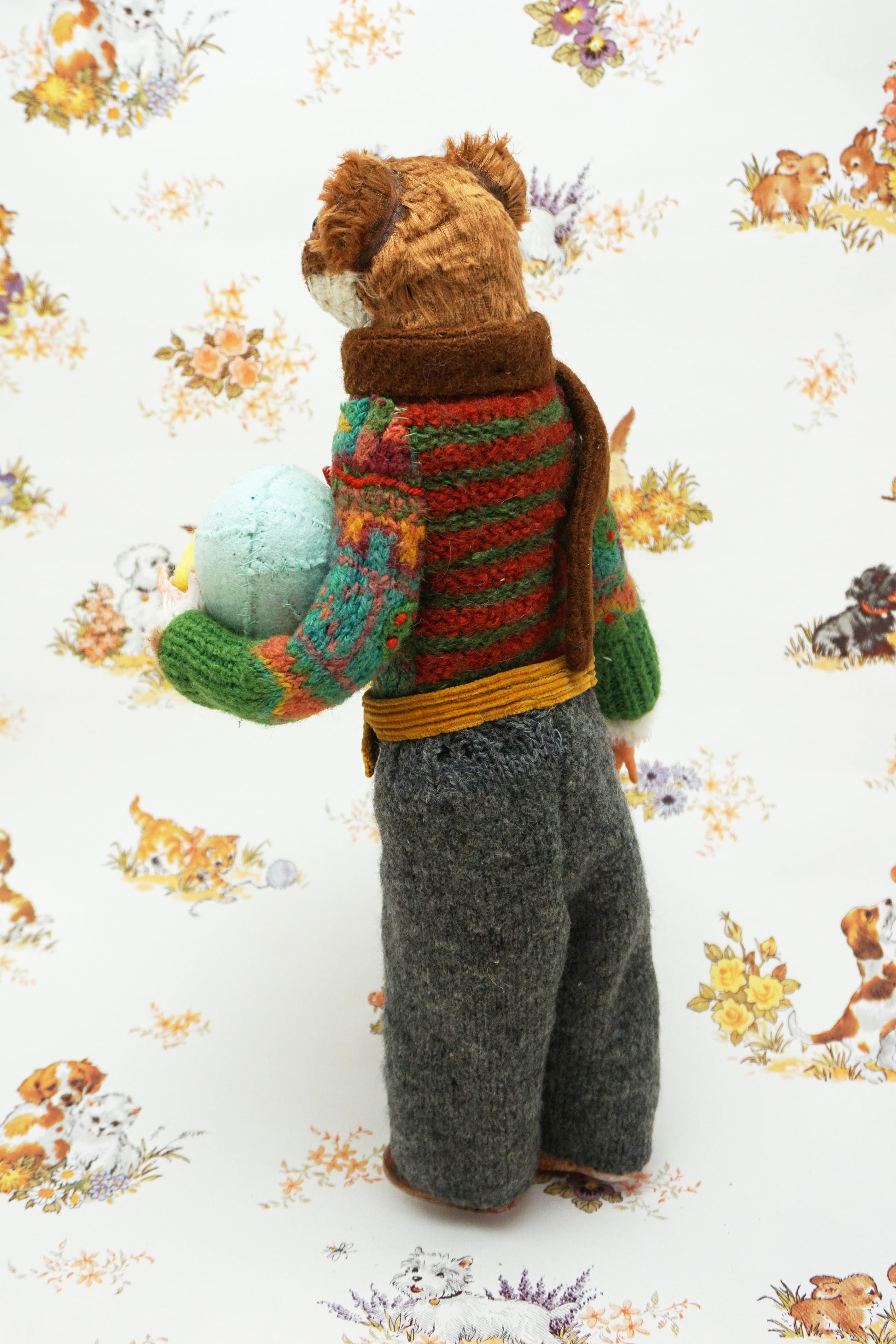 A plush weasel stands upright, wearing a green jumper, brown scarf, and grey trousers. It holds a light blue egg with yellow details. The background features a whimsical pattern of small animals and flowers.