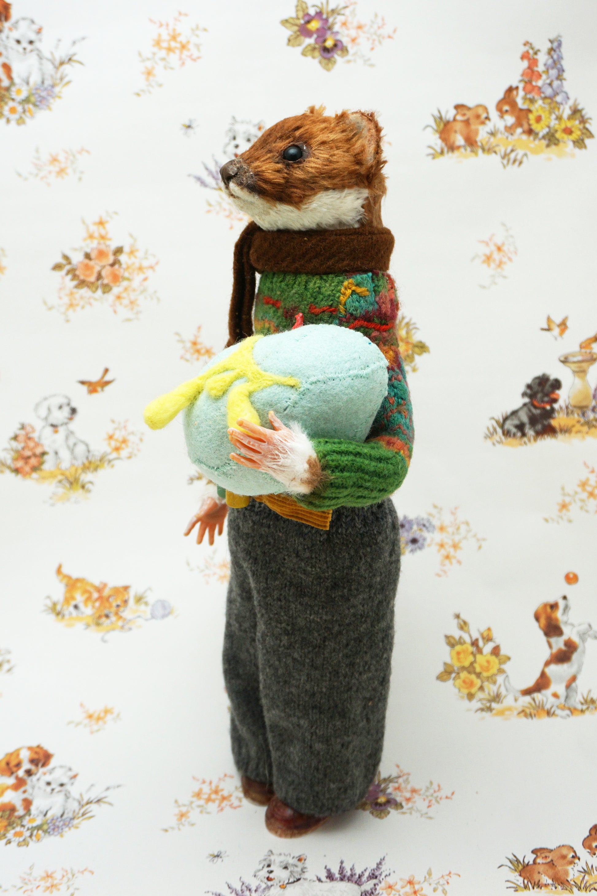 A plush weasel stands upright, wearing a green jumper, brown scarf, and grey trousers. It holds a light blue egg with yellow details. The background features a whimsical pattern of small animals and flowers.