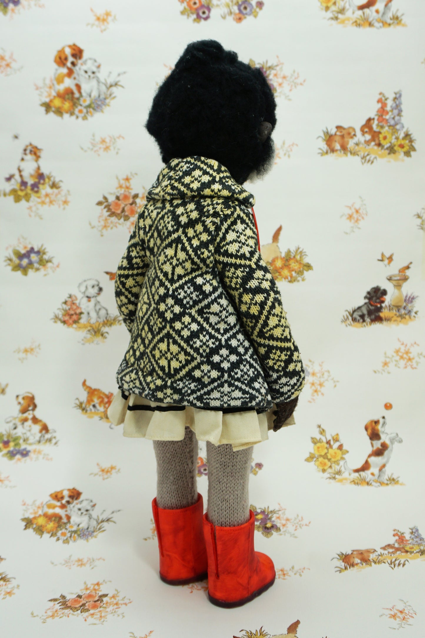A whimsical doll with a monkey-like face, wearing a patterned coat, white dress, and bright red boots. The background features a vintage-style wallpaper with illustrations of animals and flowers.