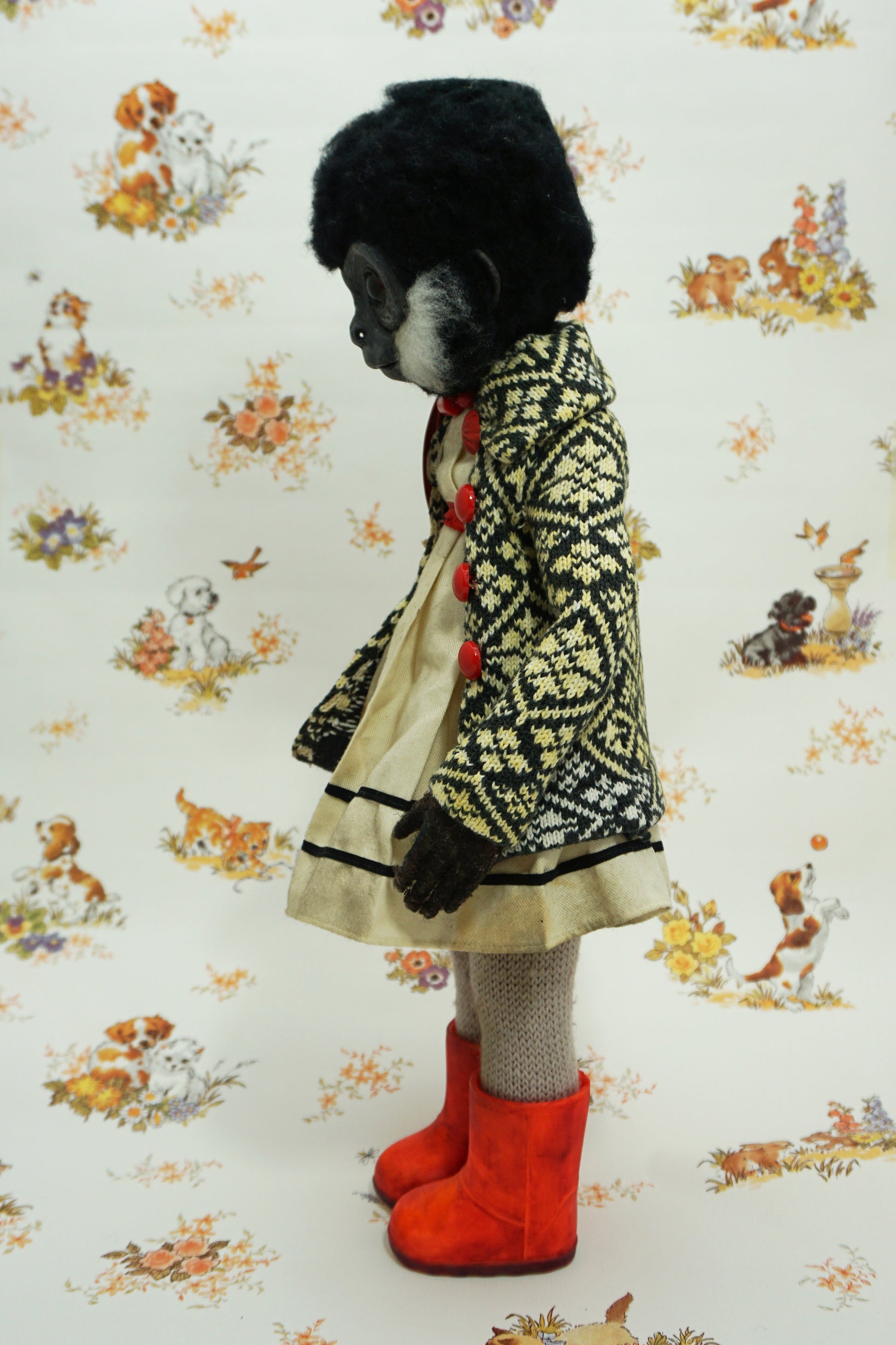 A whimsical doll with a monkey-like face, wearing a patterned coat, white dress, and bright red boots. The background features a vintage-style wallpaper with illustrations of animals and flowers.